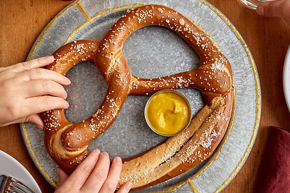 A large, salted pretzel served on a round metal plate with a small dish of yellow mustard in the center. Two hands are reaching for the pretzel, and the table is set with a red napkin, a fork and knife, a glass of water, and a beverage.