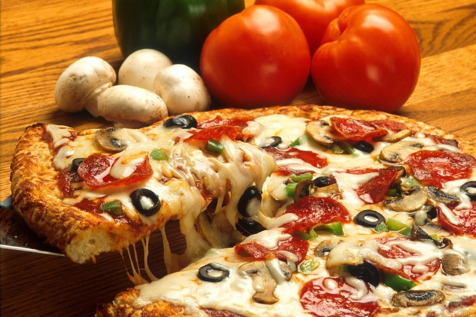 A freshly baked pizza loaded with melted cheese, pepperoni, mushrooms, black olives, and green peppers, with a slice being pulled away showing gooey cheese strands. Fresh tomatoes, mushrooms, and a green bell pepper are arranged in the background on a wooden table.